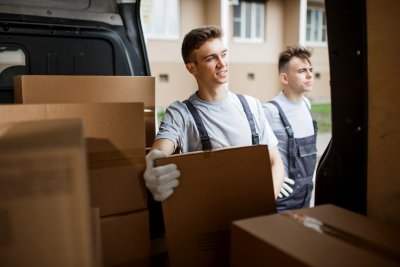 Two young handsome workers wearing uniforms are standing next to the van full of boxes. House move, mover service